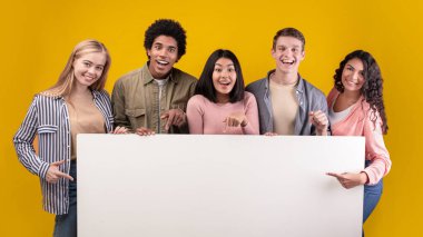 Bir grup arkadaş gülüyor, iyi ve olumlu ruh hallerini paylaşıyorlar. Ağzı açık genç çok ırklı mutlu öğrenciler boş afişi tutarlar, turuncu arka planda izole edilmiş, stüdyo çekimi, panorama