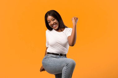 Öyle mi? Turuncu stüdyo arka planında başarıyı kutlayan güzel siyahi bayan, fotokopi alanı. Mutlu Afro-Amerikan kadını pozitif duygularını ifade ediyor, vücudunu hareket ettiriyor, dizini çekiyor ve yumruğunu kaldırıyor.