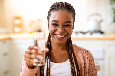 Neşeli siyahi kadın bardağı suyla tutuyor, kameraya taze içecek sunuyor, diş telleri ile gülümseyen Afrikalı Amerikalı kadın mutfak içinde sağlıklı bir sıvı kullanıyor, bedava alan sağlıyor.
