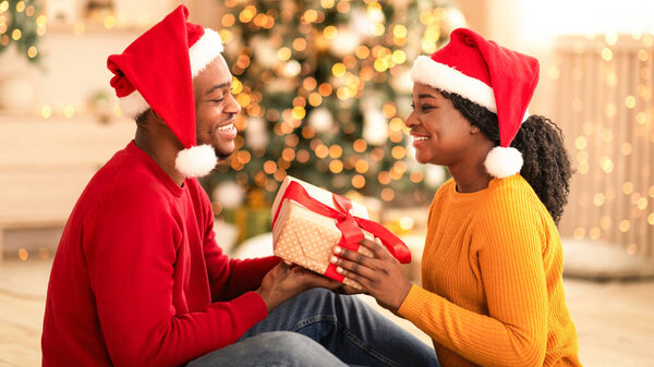 Романтика, Новый год и Рождество вместе. Счастливые молодые африканские американец мужчина и женщина в Санта-Шапки обмениваются подарками в интерьере с елкой и гостиной декор с светящимися гирляндами, профиль
