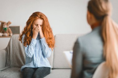 Psikolojik Danışmanlık. Çocuk psikoloğu ofiste ya da evde çalışan üzgün bir kız ağlıyor, yüzünü kapatıyor, aile sorunlarından bahsediyor, stres yaşıyor, kanepede oturuyor.