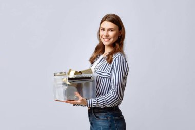 Genç bir kadın, elinde geri dönüşümlü malzemelerle dolu şeffaf bir konteynır tutarken geniş bir gülümsemeyle evin içinde duruyor. Çevre dostu uygulamalara ve sürdürülebilirliğe bağlılığını gösteriyor.