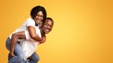 Komik siyah aşıklar. Turuncu stüdyo arka planında poz veren genç erkek ve kadın. Kopya alanı. Neşeli Afrikalı Amerikalı kadın erkeğini sırtında taşıyor, reklam için boş bir alanda duruyor.