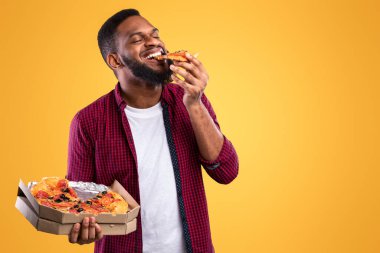 Mutlu Afrikalı genç adam pizzanın tadını çıkarıyor. Lezzetli dilimler, sarı arka planda bir kutuyla poz veriyor. Stüdyoda İtalyan pizzası yiyen abur cubur aşığı. Sağlıksız erkek beslenmesi ve aldatma yemeği