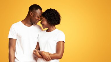 Turuncu stüdyo arka planında birbirine bağlanan, kucaklaşan ve gülümseyen kot pantolonlu, beyaz tişörtlü, çekici siyah çift ve kadının yakın çekimi. Aşk, aile, ilişkiler