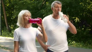 Son sınıf bir çift doğa manzaralı bir parkta koşu seansına ara veriyor. Etraflarındaki taze hava ve yeşilliğin tadını çıkarırken renkli şişelerden su yudumluyorlar..