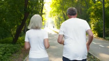 Yaşlı çiftin arka görüntüsü parkta yemyeşil bir patika boyunca yan yana koşuyor. Güneş ağaçların arasından süzülerek, egzersiz ve arkadaşlık için mükemmel huzurlu ve canlı bir atmosfer yaratır..
