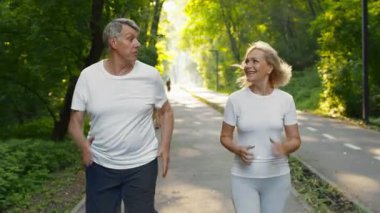 60 'lı yaşlarda bir çift ağaçlık bir patika boyunca yan yana koşuyor. Neşeli ve hareketli görünüyorlar, günün erken saatlerinde huzurlu bir parkta eğleniyorlar..