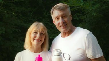 Yepyeni bir park ortamında, 60 'lı yaşlarda yaşlı bir çift ellerinde su şişeleri tutarken gülümsüyor. Koşu ve açık havada birlikte egzersiz yaparak, sağlıklarını kucaklayarak aktif bir yaşam tarzını teşvik ederler..