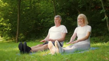 Aktif kıdemli bir çift barışçıl bir park ortamında esneme egzersizleri yapıyor. Etrafları ağaçlarla çevrili olarak, sağlıklı bir yaşam tarzını desteklerken, bu yaşlarda spor yapmanın faydalarından zevk alıyorlar..