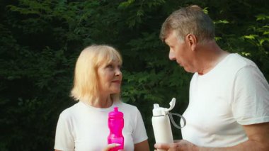 60 'lı yaşlarda yaşlı bir çift, yemyeşil bir parkta koştuktan sonra ellerinde su şişeleri tutarken birbirlerine gülümsüyor. Aktif bir yaşam tarzları var ve birlikte dışarıda vakit geçirmekten zevk alıyorlar..