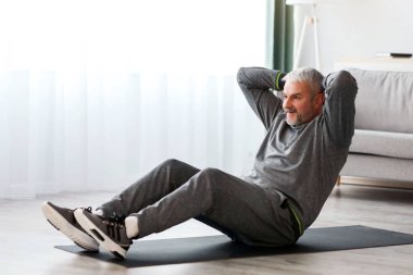 Gri spor giyimli atletik sakallı yaşlı adam evde karın kasları için egzersiz yapıyor, spor minderinde oturuyor ve çatırdıyor, fotokopi çekiyor. Sağlıklı yaşam tarzı, yaşlılar için kendini izole etme sporu