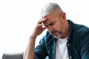 Hüsrana uğramış yaşlı sakallı adam evdeki koltukta oturuyor, kafasına dokunuyor, maddi sorunları var ya da baş ağrısı çekiyor, yakın plan, fotokopi odası olan panorama.