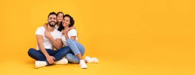 Sarı stüdyo arka planında oturan kameraya sarılıp gülümseyen üç kişilik Arap bir aile. Küçük Kızını Saran Genç Arap Ebeveynlerin Panoramik Çekimi. Metin için Boşluk