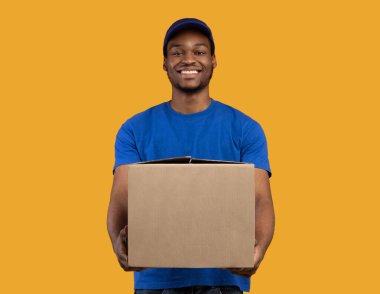 Teslimat servisi konsepti. Mavi şapkalı ve tişörtlü gülümseyen siyah postacı karton kutu tutuyor, kameraya gösteriyor ve ona veriyor, kopyalama alanı. Turuncu stüdyo arka planında duran postacı.