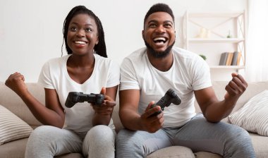 Çiftler için eğlenceli hobiler. Neşeli siyah adam ve kız video oyununda zaferi kutluyorlar, joystickleri tutuyorlar, yumruklarını kaldırıyorlar ve gülüyorlar, panorama
