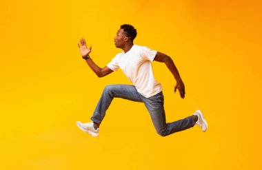Afrikalı adam atlıyor, hızlı koşuyor, satışa ya da indirime gidiyor, sarı arka planda günlük kıyafet ayakkabıları giyiyor.