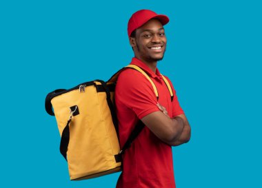 Teslimat Servisi. Kırmızı üniformalı ve termo çantalı özgüvenli siyah erkek kurye portresi katlanmış kollarla poz veriyor ve mavi stüdyo arka planında izole edilmiş kameraya bakıyor.