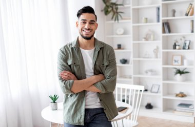 Yakışıklı genç bir Arap 'ın portresi evde kollarını kavuşturmuş, gülümseyen orta doğulu bir adam aydınlık odada rahat kıyafetler giymiş, kameraya gülümsüyor.