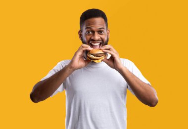 Sarı arka planda duran kameraya gülümseyen ve hamburger yiyen neşeli, aç Afrikalı adam. Stüdyo çekimi. Abur cubur yiyici çizburgerin tadını çıkarıyor. Fast food ve Hile Yemekleri