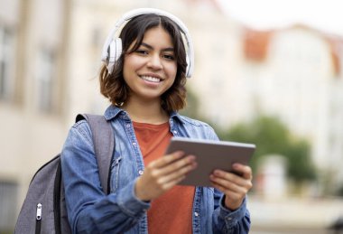 Kablosuz kulaklıklı gülümseyen Arap kız öğrencinin portresi açık havada dijital tablet kullanıyor sırt çantalı neşeli Orta Doğulu kadın üniversite dersinden sonra modern Gadget ile dinleniyor.