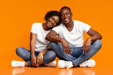 Güzel Afro-Amerikan çift yerde oturmuş, birbirlerine sarılıyorlar, kameraya bakıyorlar ve gülümsüyorlar, boş bir alan. Genç mutlu siyah adam ve kadın turuncu stüdyo arka planında birlikte poz veriyorlar.