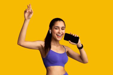 Müzik aşığı. Telsiz kulaklık takan, akıllı telefon tutan, mikrofon takan ve şarkı söyleyen, turuncu stüdyoda izole edilmiş favori şarkı listesini dinleyen neşeli ve mutlu bir kadının portresi.