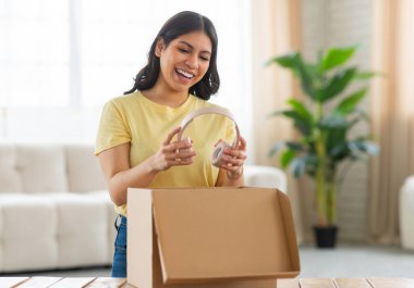 Genç bir ortadoğulu kadın, elinde kulaklıklarla masaya bir karton kutu yerleştirirken heyacan ve parlak bir gülümseme ile güneşli oturma odasında duruyor.