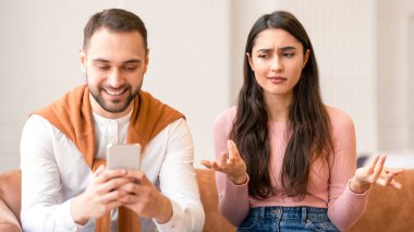 Kafası karışmış Orta Doğulu bir kadın, akıllı telefondan kocaya bakarken evde koltukta oturan sadakatsizlikten şüpheleniyor. Arap Adam Karısını Görmezden Gelen Cep Telefonu Kullanıyor. İlişki Sorunları