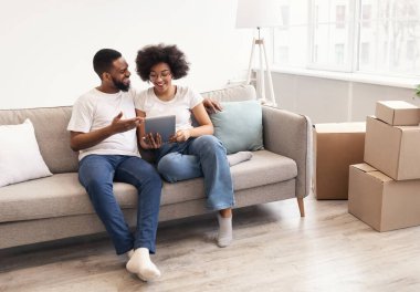 Yeni dairede taşındıktan sonra taşınan koltuklarda oturan mutlu siyah eşler tabletini kullanıyor. İnternette arama yapan çiftler taşınma işlemine hazırlanıyor. Boşluğu Kopyala