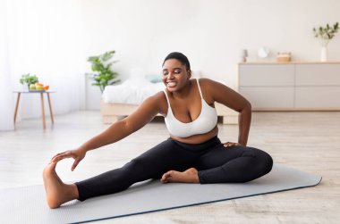 Kıvrımlı siyah kadın bacaklarını açıyor, yoga minderi üzerinde egzersiz yapıyor, aktif bir hayat sürüyor, evde kilo vermeye çalışıyor. Ayrıca zayıf düşürme programı sırasında spor yapan Afrikalı Amerikalı bir kadın.