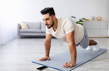 Güç egzersizi konsepti. Genç bir Arap 'ın evde şınav çekmesi ya da kalas çekmesi, akıllı telefondan video izlemesi, boş alan. Atletik Doğulu bir adam covid-19 tecridi sırasında aile içi eğitim alıyor.