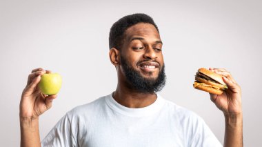 Sağlıklı ve Sağlıksız Yiyecekler. Aç Afrikalı Adam, Burger ve Apple 'ın stüdyoda durması arasında seçim yapıyor. Diyet yemek, erkek beslenmesi ve abur cubur konsepti.