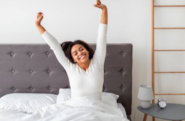 Neşeli pozitif siyah bayan uyandıktan sonra kollarını açıp geri dönüyor. Mutlu güzel Afrikalı Afrikalı Amerikalı kadın yatakta oturuyor, harika hissediyor, günün iyi başlangıcının tadını çıkarıyor, bedava fotokopi alanı