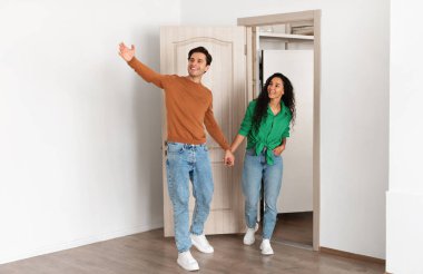 Look At This. Portrait of excited guy inviting lady to enter home, welcoming to living room, happy smiling man holding womans hand showing new modern apartment, walking in flat together