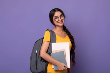 Eğitim Konsepti. Gözlüklü, elinde not defterleriyle kameraya bakan gülümseyen Hintli kız öğrencinin portresi. Sırt çantalı bir kadın mor stüdyo arka planında yalnız poz veriyor.