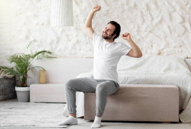 İyi sabahlar. Mutlu sakallı genç adam yatakta uyanıyor ve kollarını açıyor, gözleri kapalı gülümsüyor, pijama ve çorap giyiyor, fotokopi odası olan bir panorama, yatak odası.