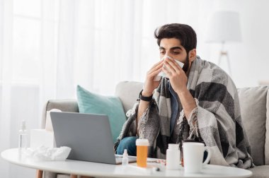 Sıcak battaniyeye sarılmış, dizüstü bilgisayara bakan, kağıt mendile hapşıran, evdeki kanepede oturan, boş alana bakan Arap bir adam. Sinüzit hastalığı, soğuk algınlığı ve grip belirtileri