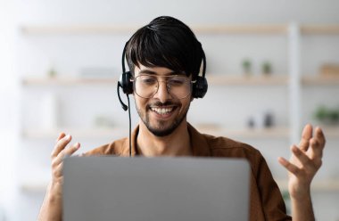 Gözlüklü gülümseyen genç Arap adamın portresi meslektaşları veya müşterileriyle görüntülü konuşma yapıyor, bilgisayar ekranına bakıyor ve el kol hareketi yapıyor, kulaklık kullanıyor, proje yöneticisi çevrimiçi iş konferansı yapıyor.