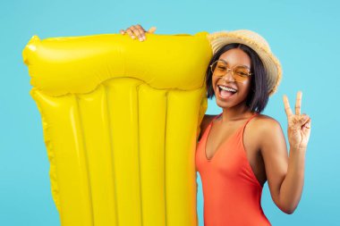 Yaz tatili dinlenme zamanı. Sarı lilo tutan mayolu neşeli siyah bayan ve mavi stüdyo arka planında barış jesti gösteriyor. Kaygısız Afrikalı Amerikalı kadın yaz tatilinin tadını çıkarıyor.