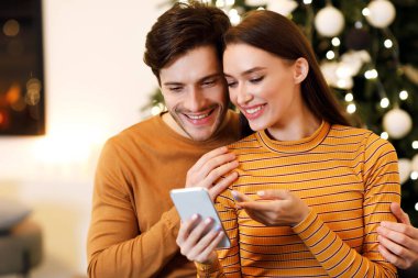 Neşeli bir milenyum çifti cep telefonuyla birlikte ne alacaklarını seçiyorlar, evdeki koltukta oturan aleti paylaşıyorlar, mutlu adam ekranı işaret eden kadını kucaklıyor, konuşuyor ve seçenekleri tartışıyorlar.