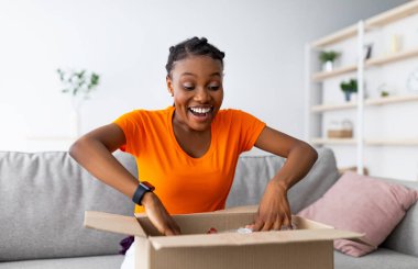 Duygusal siyahi kadın müşteri uzun zamandır beklenen paketi alıyor, karton kutuyu evde açıyor, büyük alışverişlerle tatmin oluyor. Çevrimiçi alışveriş ve dağıtım kavramı