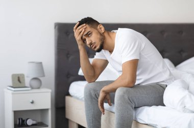 Apathy Concept. Portrait Of Depressed Young Middle Eastern Man Sitting On Bed In Bedroom, Upset Millennial Arab Guy Touching Head And Looking Away With Sorrow, Suffering Seasonal Depression, Closeup