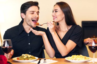 Romantic Date Concept. Smiling beautiful girlfriend feeding handsome guy with pasta. Portrait of happy excited family sitting at table and eating meal at restaurant or at home, drinking red wine