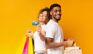 Dost memnun Afro-Amerikan erkek ve kadın ile kredi kartı ve alışveriş torbaları, ayakta arka arkaya üzerinde turuncu arka plan, panorama