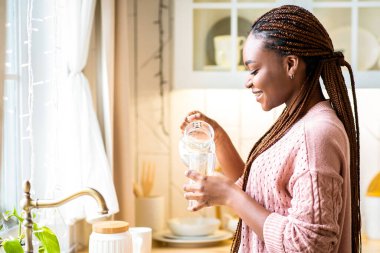 Genç Afrikalı Amerikalı bayan mutfaktaki sürahi bardaktan su dolduruyor. Saçları örgülü, sırıtan, ferahlatıcı içecekten keyif alan, vücut suyuyla ilgilenen, boş alanla yan görünüşlü siyah bir kadın.