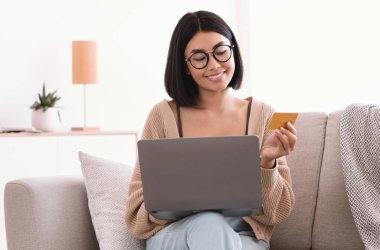 İnternetten Evden Alışveriş, Ticaret Konsepti. Gülümseyen Asyalı genç kadın evdeki koltukta oturuyor, internetten kıyafet ve ürün sipariş ediyor, kredi kartıyla ödüyor ve dizüstü bilgisayar kullanıyor.