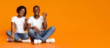 Yerde oturan mutlu siyah aşıklar, parmaklarıyla fotokopi odasına sarılıp işaret ediyorlar, turuncu stüdyo arka planı, panoramik fotoğraf. Gülümseyen Afro-Amerikan kadın reklamlara nişan alıyor.