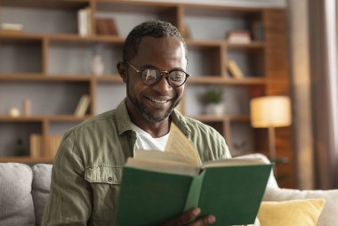 Oturma odasının iç kısmında gözlüklü, neşeli, orta yaşlı Afro-Amerikan erkek portresi. Yaklaş. Boş zamanlarında edebiyat, öğrenim ve bilgi, yeni çok satanlar, hobi ve evde dinlenme.