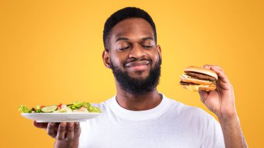 Sağlıklı vs. Sağlıksız. Aç Afrikalı Amerikalı Adam, Burger ve Salata Pozu arasında seçim yapıyor. Sarı Stüdyo Arkaplanda duruyor. Diyet, Beslenme ve Gıda Seçimi Konseptinde Aldatma